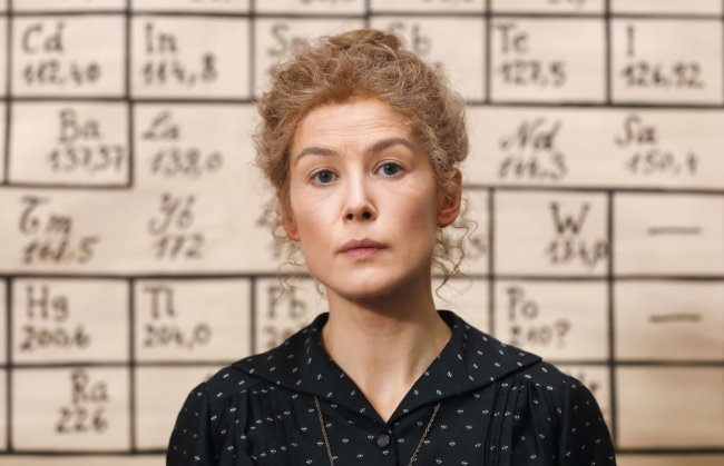 Foto de divulgação do filme Radioactive, protagonizado por Rosamund Pike; a atriz está caracterizada em frente a uma lousa preenchida com elementos químicos