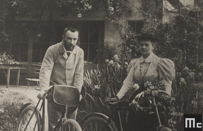 Foto em preto e branco da cientista Marie Curie andando de bicicleta com seu marido, Pierre, em Paris; eles posam para a foto em uma vilinha florida