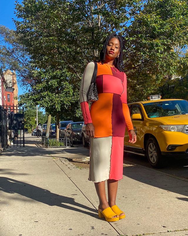 Garota usando vestido midi de manga longa de patchwork nas cores laranja, bordô, vermelho e bege. Ela está com um tamanco amarelo e uma bolsa de estampa de zebrinha pendurada em um dos ombros. Ela está em pé, olhando em direção à câmera, que tira a foto de cima para baixo.