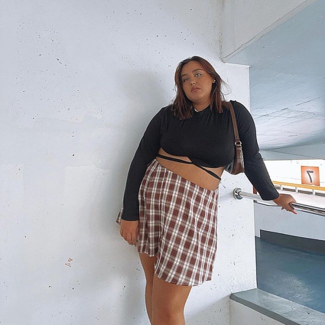 Garota está usando blusa de manga longa cropped preta com saia xadrez marrom e off-white e uma bolsa baguete pendurada em um dos ombros. Ela está com o corpo inclinado e uma das mãos apoiadas em um corrimão.