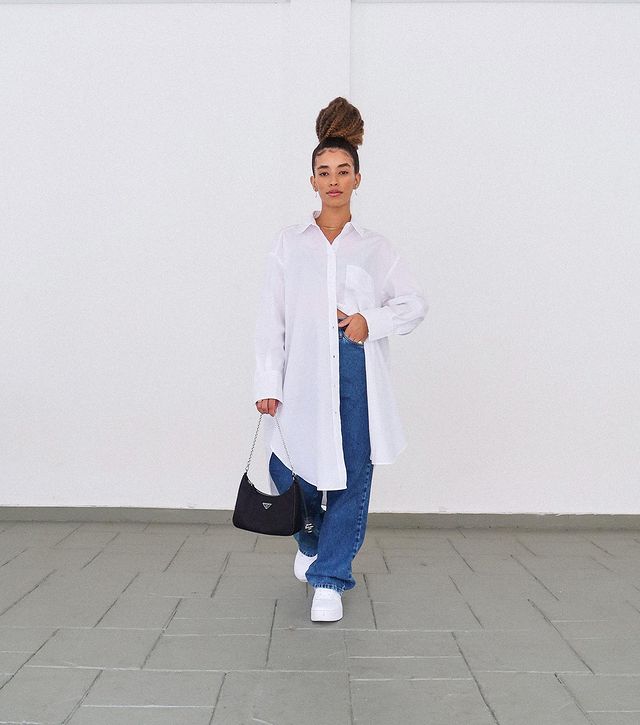 Mulher está usando camisa branca oversized com calça jeans baggy, tênis branco e bolsa preta em uma das mãos. Ela está em pé, os cabelos presos em um coque e uma das mãos na cintura.