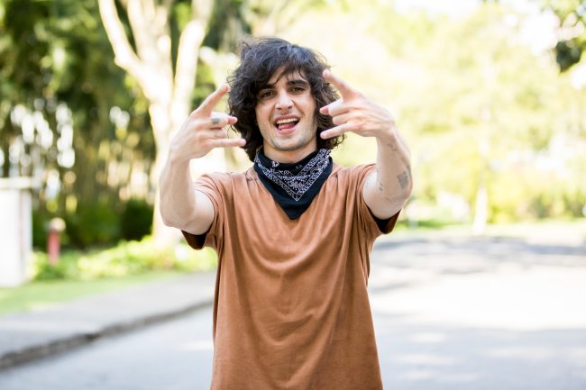 BBB21 Fiuk posando para foto com as mãos fazendo símbolo do rock usando camiseta marrom e bandana preta amarrada no pescoço