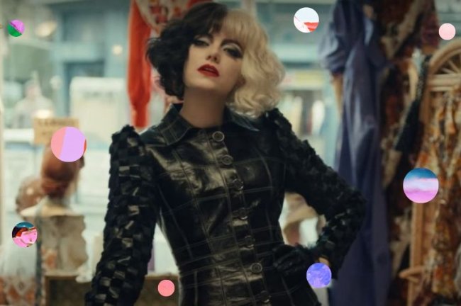 Emma Stone no filme Cruella, vestida como a personagem, usando peruca de cabelo na cor preta e branca, jaqueta de couro preta, com a mão na cintura com expressão séria.