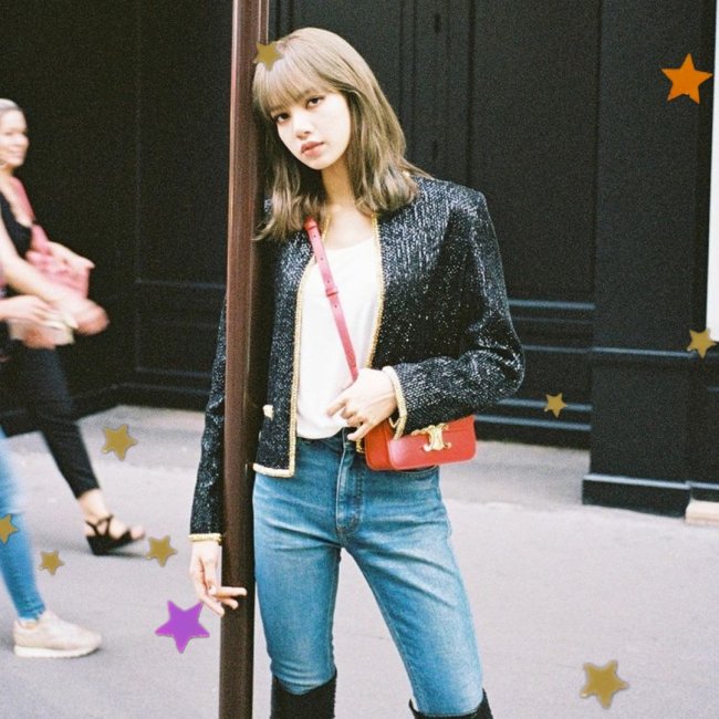 Lisa, integrante do grupo Blackpink, posando para foto encostada em um ferro, usando blazer preto, bolsa vermelha, calça jeans e bota preta. Sua expressão é séria.