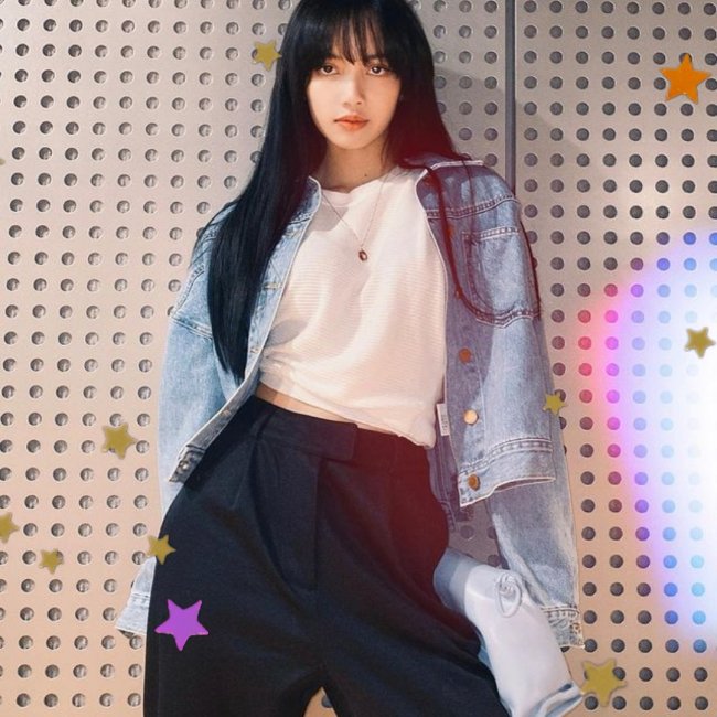 Lisa, integrante do grupo Blackpink, encostada na parede com aparência séria, com o cabelo escurto, usando jaqueta jeans clara, blusa branca e calça azul.