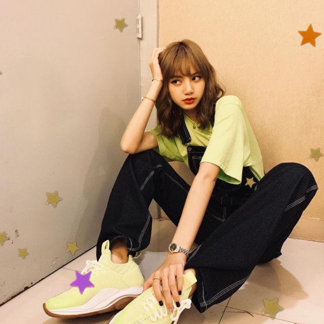 Lisa, integrante do grupo Blackpink, sentada com uma das mãos tocando seu pé e a outra apoiada na cabeça; com uma expressão pensativa;usando camiseta verde clara, assim como seu tênis e calça jeans.