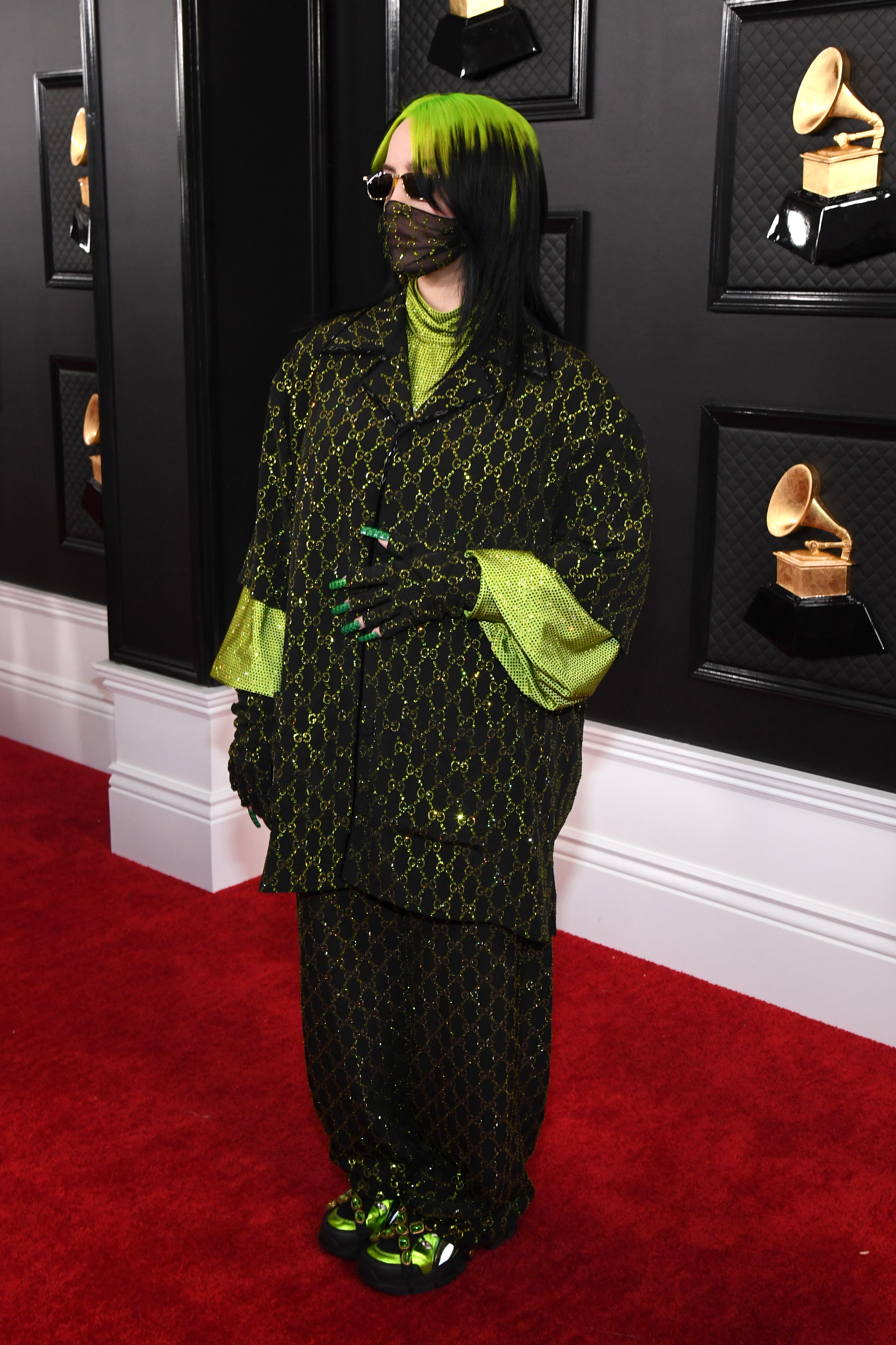Billie Eilish no tapete vermelho do Grammy Awards Foto da cantora Billie Eilish no tapete vermelho do Grammy Awards. Ela usa uma camisa oversized preta com detalhes verdes, calça preta com detalhes verdes e tênis preto com detalhes verdes. Ela não olha para a câmera.