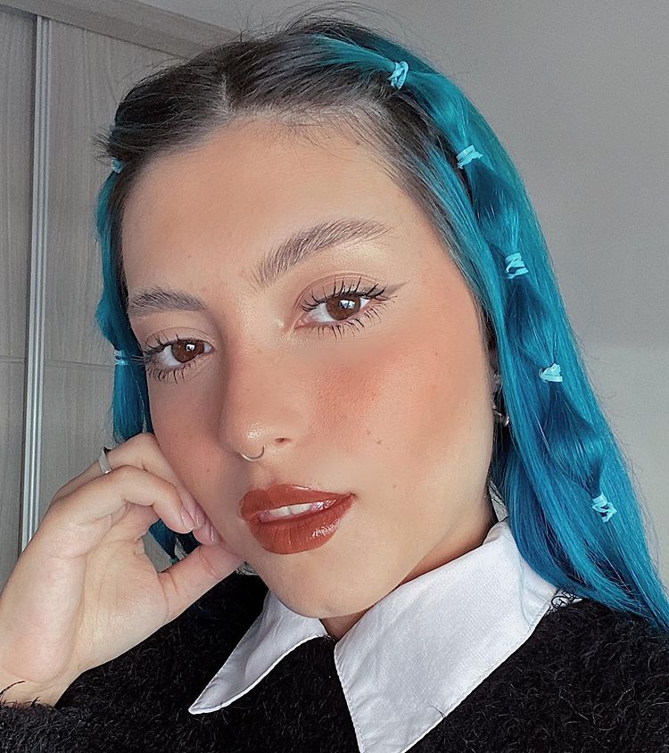 Selfie de uma mulher. Ela está usando uma camisa branca com um casaco de sobreposição, cabelo azul com a franja presa com elásticos coloridos. Ela olha para a câmera, não sorri e apoia o rosto na mão direita.