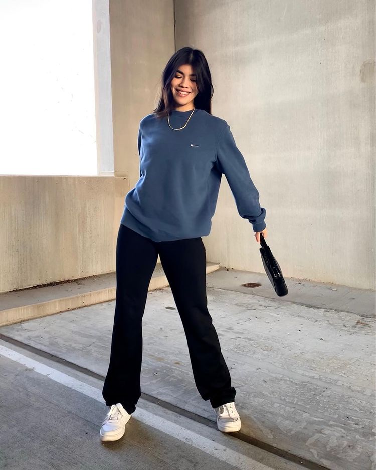 Foto de uma mulher no estacionamento. Ela usa um moletom azul, calça legging bailarina, tênis branco e shoulder bag preta. Ela olha para o chão e sorri.