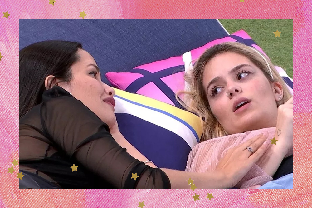 Foto de Juliette e Viih Tube deitadas no jardim do BBB21 enquanto conversam; Juliette usa roupa preta e Viih Tube usa roupa rosa; Juliette está com a mão se movendo para segurar na mão da influenciadora e as duas se olham; a moldura da imagem é uma textura de tinta em tons de rosa e estrelas amarelas são usadas como decoração