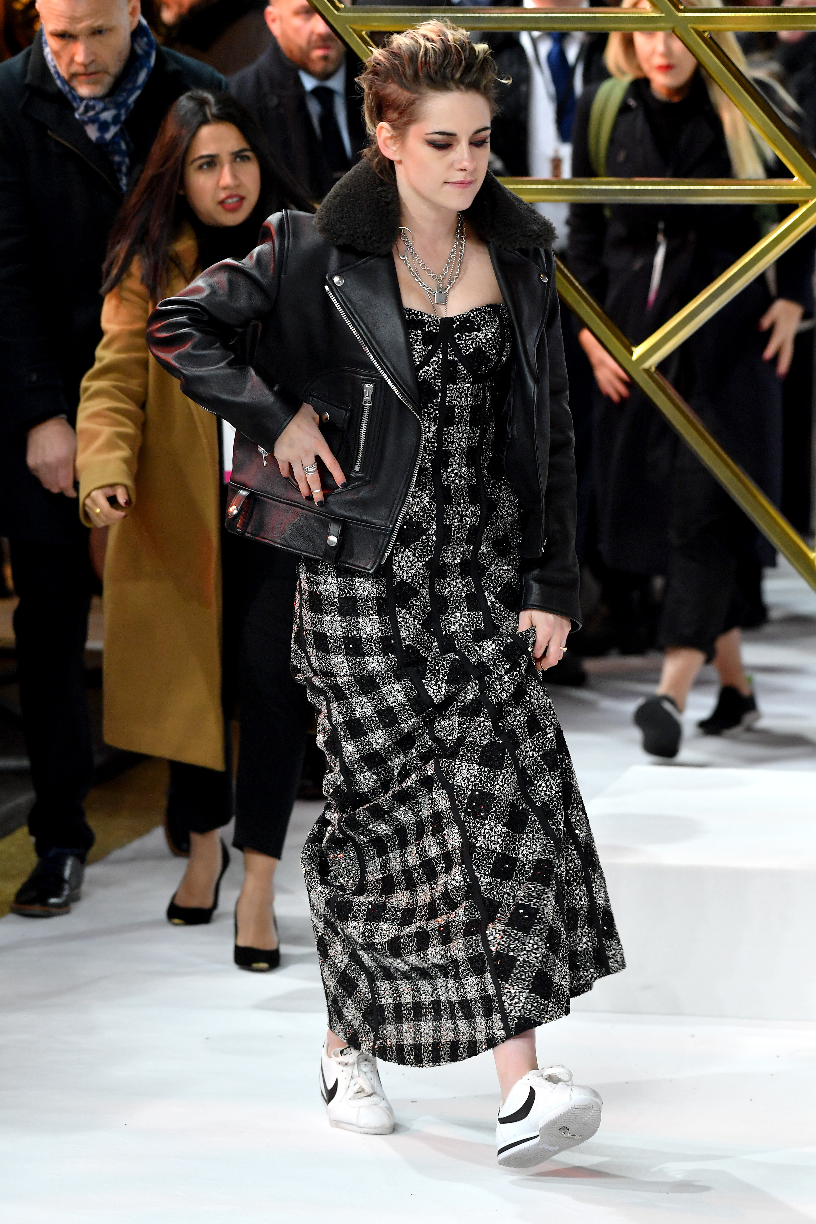 Kristen Stewart de tênis no tapete vermelho Kristen Stewart no tapete vermelho da premiere do filme As Panteras em 2019. Ela está usando um vestido com estampa xadrez preta e cinza, jaqueta de couro preta, colares de corrente prata e tênis. Ela caminha e não olha para a câmera.