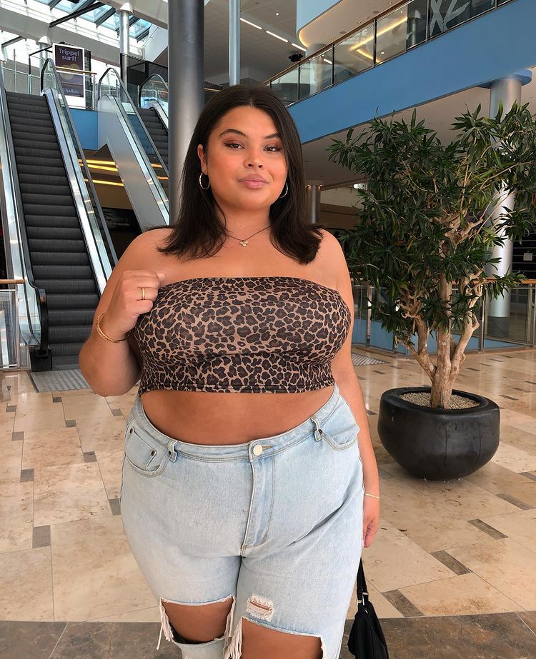 look-blusa-animal-print-joannapincerato Garota usando top cropped de animal print com calça jeans e segurando uma bolsa preta. Ela está em um shopping, olhando pra frente com expressão facial séria.