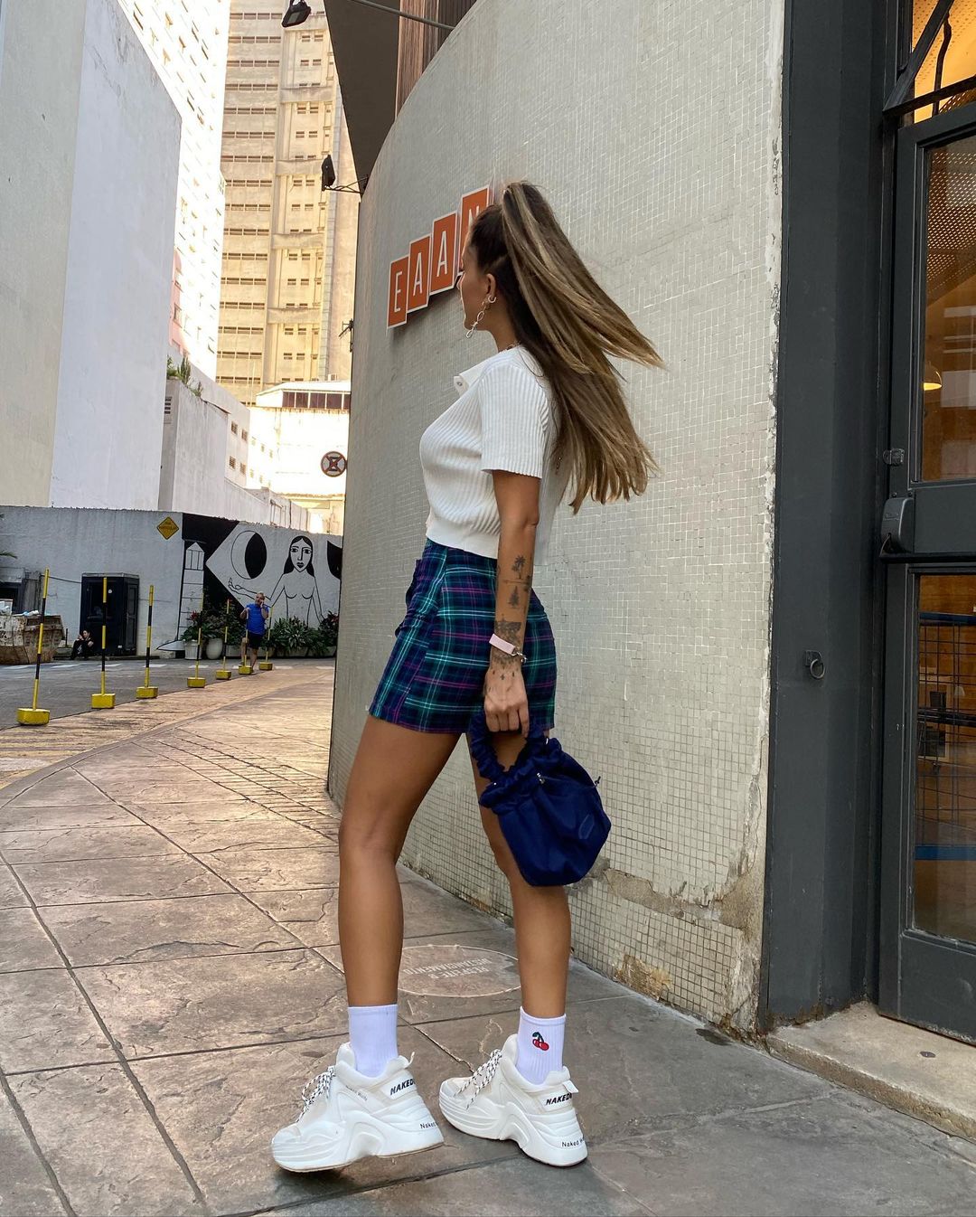look-saia-xadrez-tenis-branco A mulher está em uma rua. Ela veste uma camiseta polo branca, saia azul com estampa xadrez e um tênis plataforma branco. Ela está de perfil para a foto.
