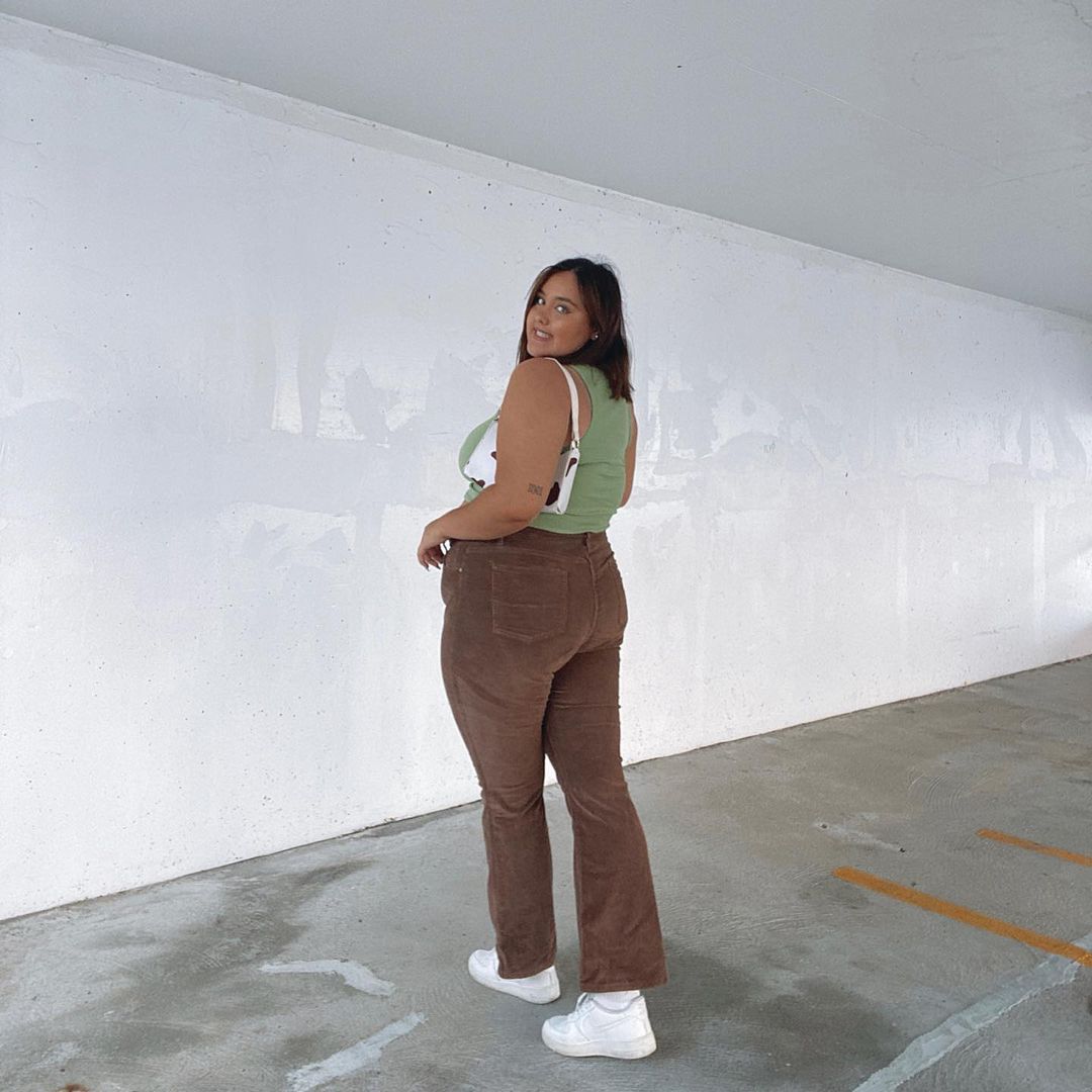 look-tenis-branco-calca-marrom A mulher está em um estacionamento. Ela veste um cropped verde menta, calça marrom, bolsa baguete com estampa de vaquinha, tênis branco e está com o cabelo solto. Ela olha para a câmera sorrindo.