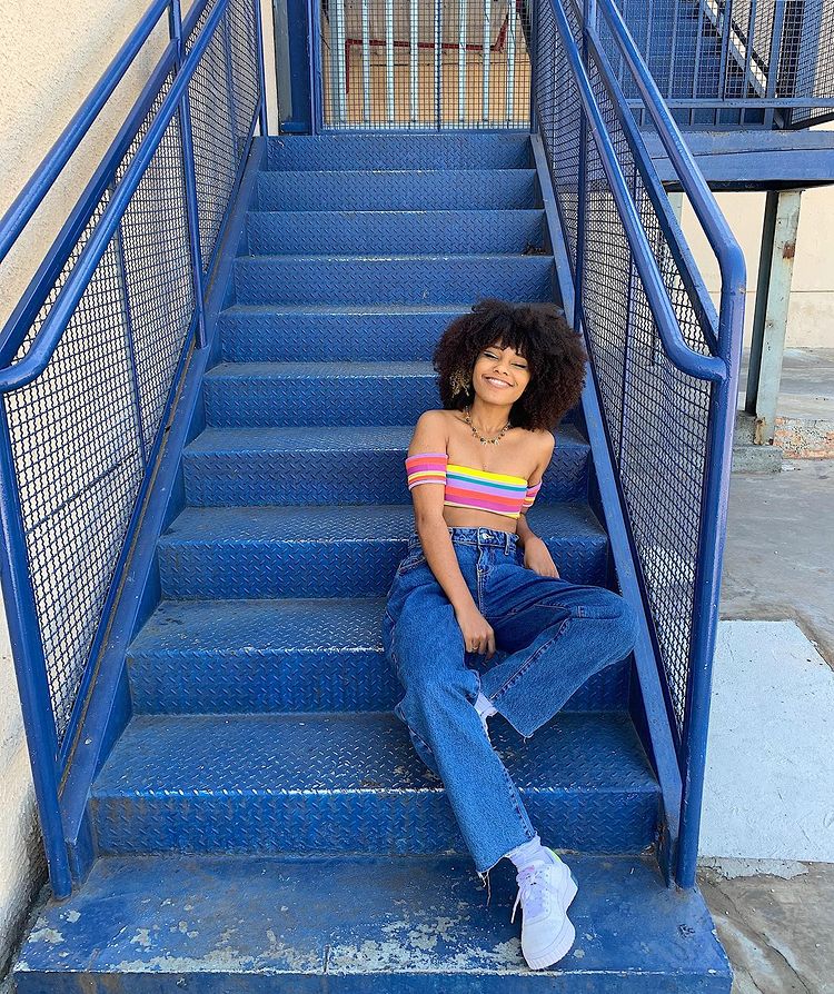 look-tenis-branco-wide-leg Mulher sentada em uma escada azul. Ela usa um cropped listrado e colorido, calça wide leg azul, um tênis branco e o cabelo crespo solto. Ela olha para a câmera sorrindo.