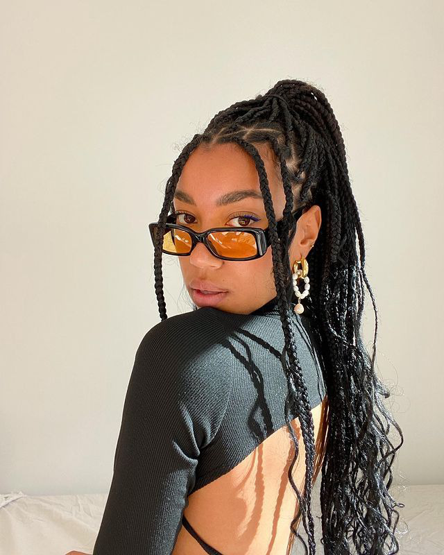 oculos-de-sol-tendencia-amaka.hamelijnck Garota usando blusa preta com decote nas costas e óculos de sol de armação preta com lentes alaranjadas e retangulares. Ela está olhando por cima do ombro com expressão facial séria.
