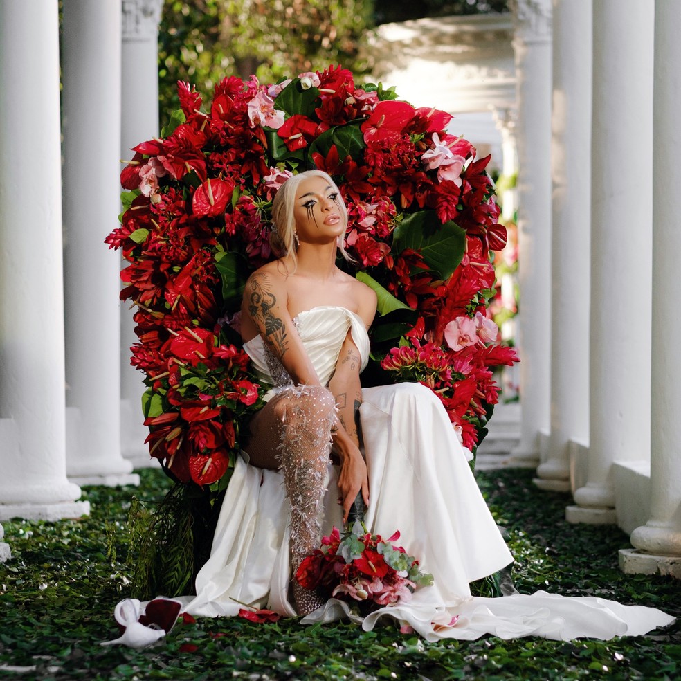 Pabllo Vittar sentada em cadeira decorada com rosas vermelhas, ela usa um vestido de noiva branco e segura um buquê de flores; a maquiagem da cantora é escura e seu rímel está borrado, indicando que ela teria chorado