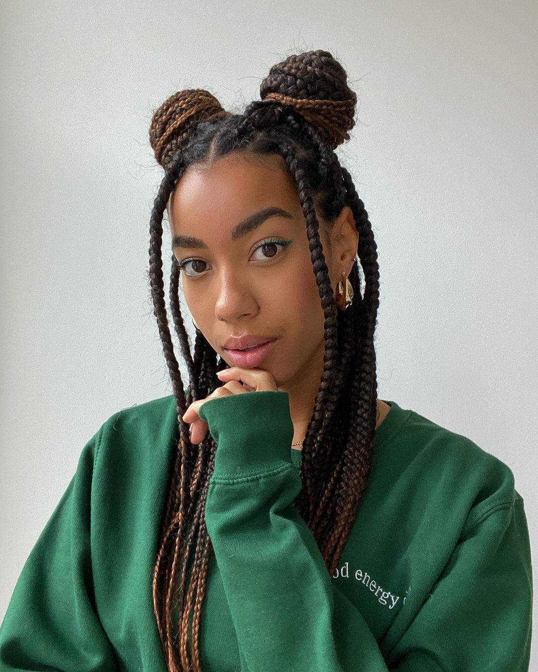 penteado_coque_duplo_tranca Selfie de uma mulher. Ela veste um moletom verde e cabelo trançado solto com coque dupo. Ela olha para a câmera e apoia a mão esquerda no queixo.