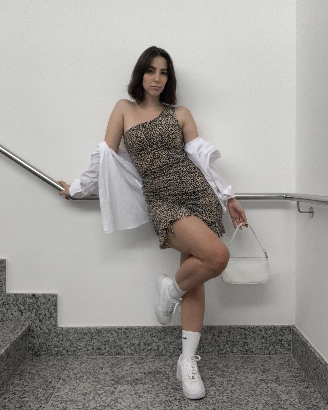vestido-animal-print-isabelabaccarini Garota usando vestido de um ombro só com estampa de oncinha, camisa branca por cima caída nos ombros, tênis branco e meia branca. Uma das mãos está apoiada no corrimão e a outra está segurando uma bolsa branca. Ela está olhando para frente com expressão facial séria.