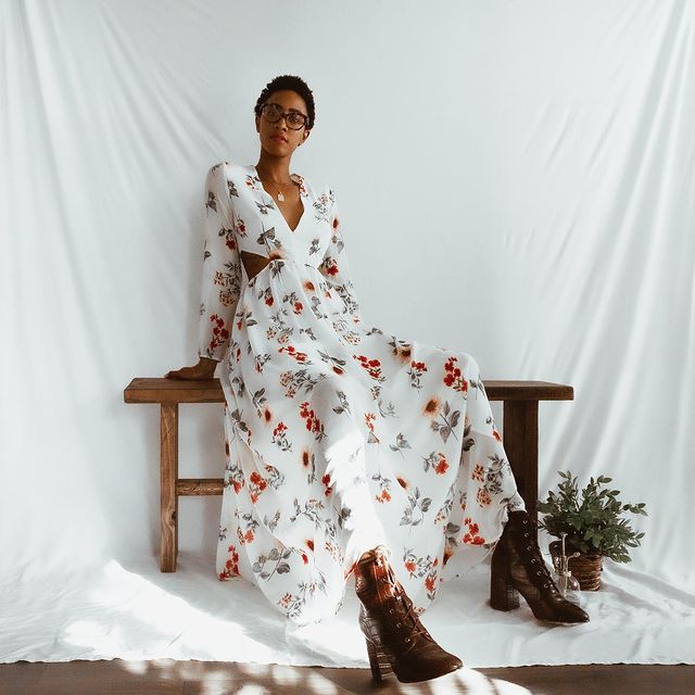 Garota usando vestido longo floral com fundo branco e botinha marrom com salto e bico fino de verniz. Ela está sentada em um banquinho de madeira, com as duas mãos para trás do corpo. Ao fundo, há um pano branco que serve como cenário para a foto.