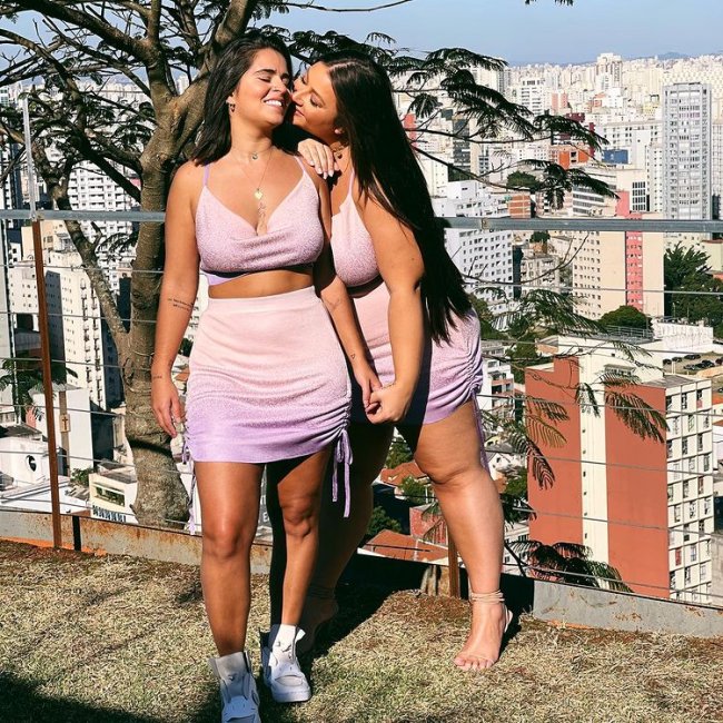 Casal posando com uma jovem de frente e a outra de lado a beijando, elas usam a roupa igual. Um top e uma saia rosa claro. Uma usa tênis e a outra uma sandalia de salto. As duas estão com uma expressão alegre.