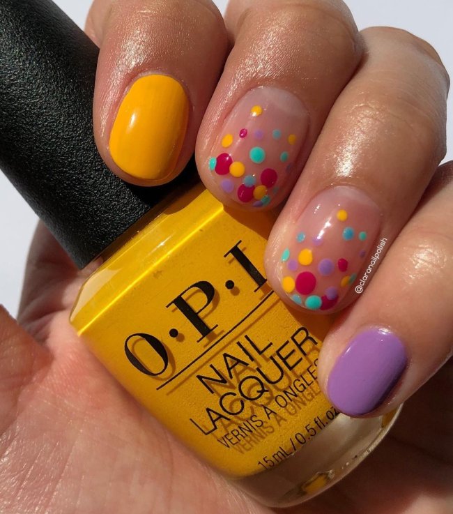 Foto com destaque nas unhas com nail art de bolinha, dessa vez com algumas unhas lilás, outras amarelas, e outras com bolinhas nas cores lilás, vermelho, amarelo e verde.