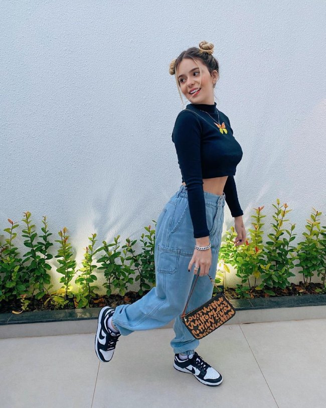 Viih tube com calça jogger jeans posando com o pé esquerdo levantado, segurando uma bolsa baguete e usando cropped preto, a influenciadora aparece sorridente.