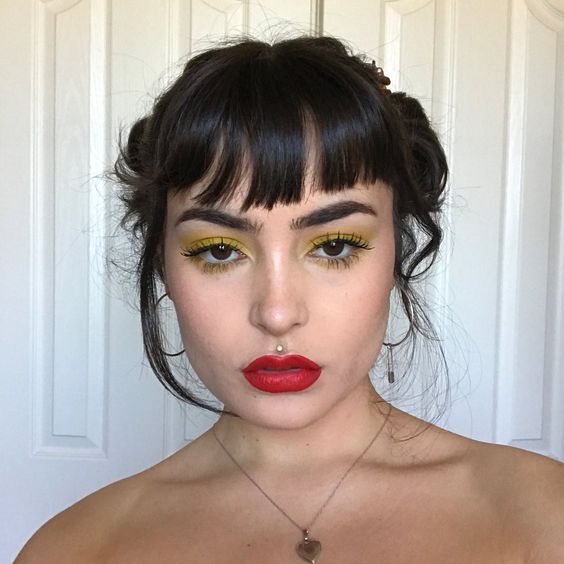 baby bangs Selfie de uma mulher. Ela usa um colar com pingente de coração, cabelo preso em dois coques laterais, franja curtinha solta e batom vermelho. Ela olha para a câmera e não sorri.