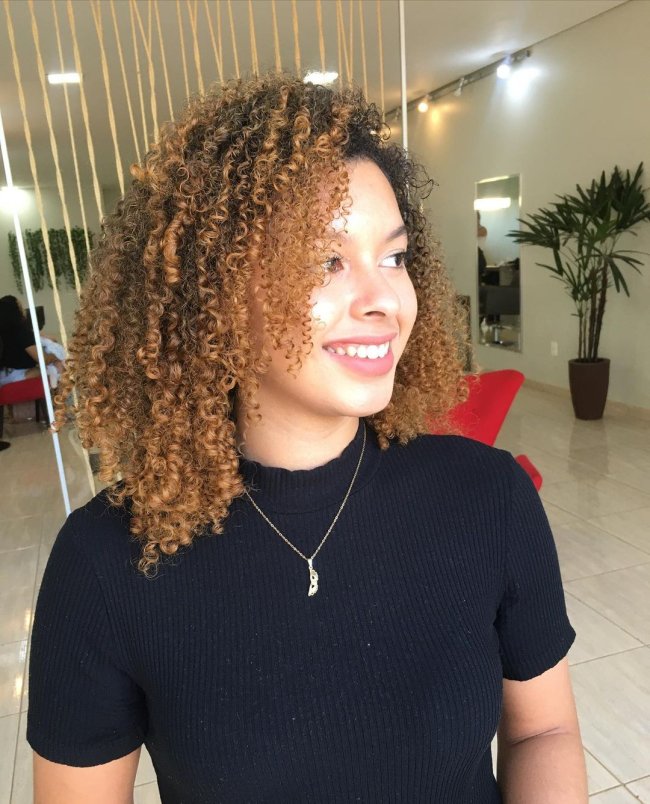 Jovem sorridente olhando para o lado, posando em salão de cabeleireiro usando camiseta preta de gola alta com colar. Ainda na imagem conseguimos ver alguns detalhes do salão.