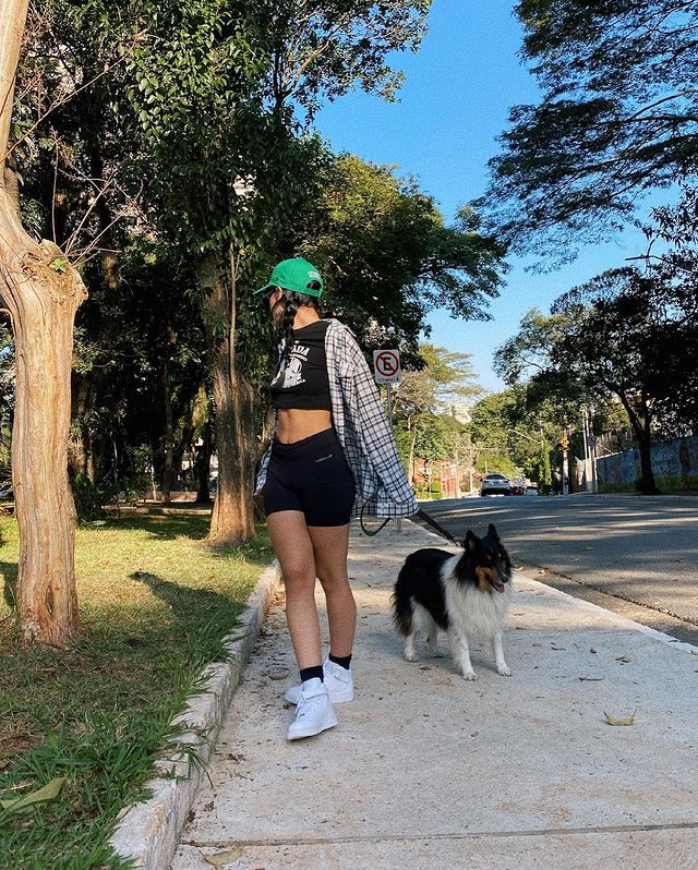 Foto de uma mulher em uma calçada com um cachorro. Ela usa uma camiseta preta, shorts preto, camisa xadrez, boné verde e tênis branco. Ela olha para trás.