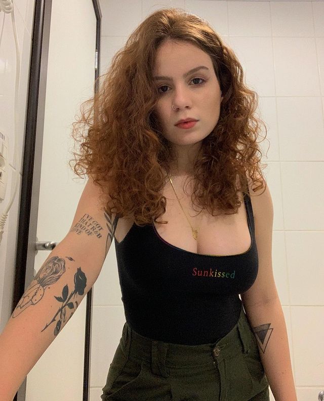 Foto de uma mulher no banheiro. Ela usa um body preto, uma calça verde militar e está com o cabelo solto. Ela olha para a câmera e não sorri.