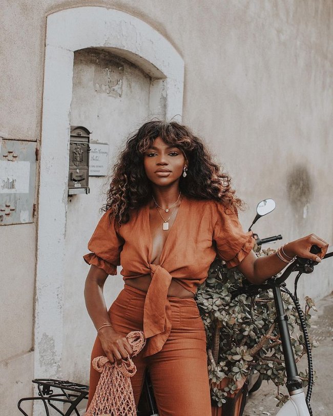 Jovem posando com look laranja monocromático, em apoiada em uma bicicleta com flores, ainda conseguimos ver uma porta branca atrás dela.