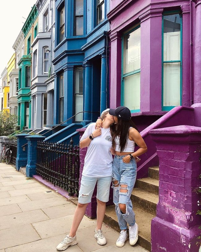 Um casal se beijando em uma rua com casas coloridas nas cores verde, cinza, azul e roxo. No casal uma moça veste camiseta branca e short, e a outra boné preto, cropped branco e calça destroyed. Não conseguimos ver o rosto delas.