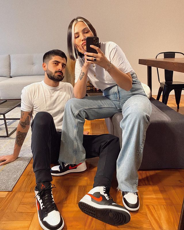 Casal posando em frente a espelho, o homem está sentado no chão usando camiseta branca e calça jeans escura, a menina está sentada em um pufe usando camiseta branca e calça jeans. Os dois estão com o mesmo tênis.