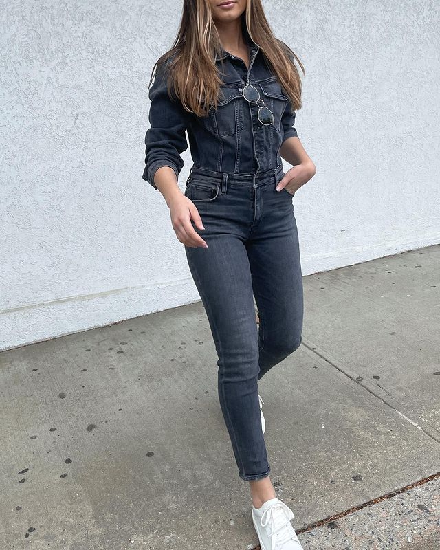 Garota usando macacão jeans em lavagem escura com tênis branco e um óculos de sol pendurado na parte de cima da roupa. A foto está cortada na altura da boca e ela está andando em direção à câmera.