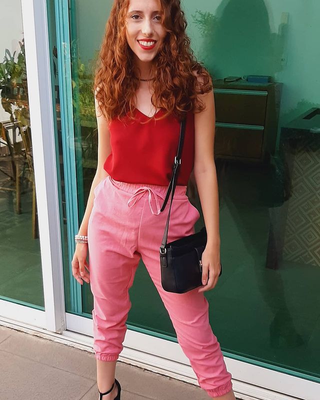 Jovem posando com regata vermelha, calça rosa e bolsa preta. Sua expressão é sorridente.