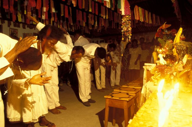 Imagem mostra um culto de candomblé realizado na Bahia. Negros vestidos de brancos se curvam em frente a estátuas de santos.