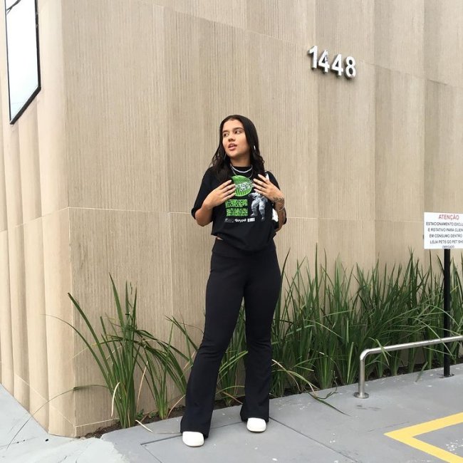 Jovem posando em frente a parede bege e detalhe de plantas na parte inferior, ela usa camiseta preta com detalhes em verde e calça bailarina. Ela olha para o lado com uma expressão séria.