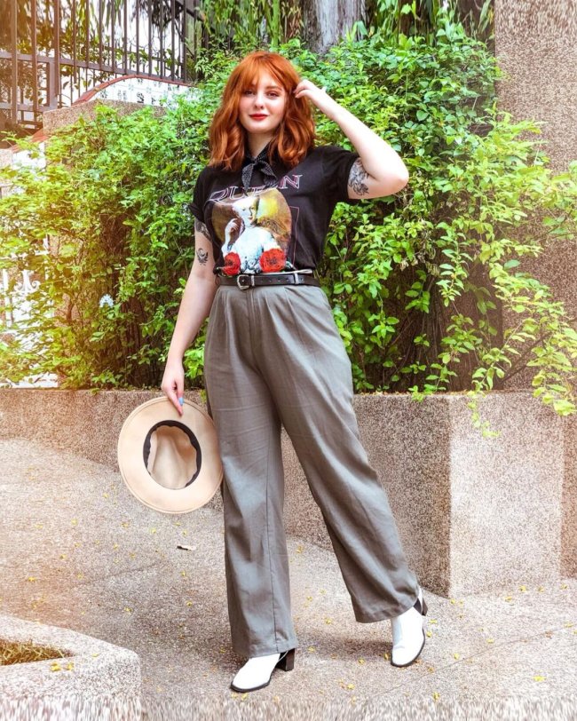 Jovem posando segurando um chapéu, com camiseta de banda e expressão sorridente.