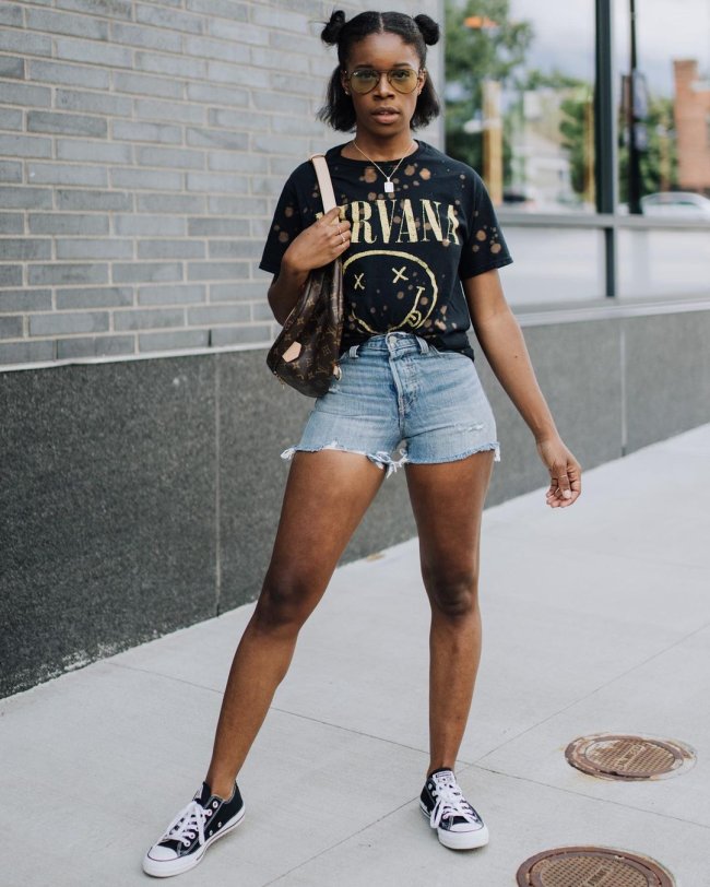 Jovem posando com short jeans e camiseta de banda. Sua expressão é séria e ela usa o penteado space buns em seus cabelos.