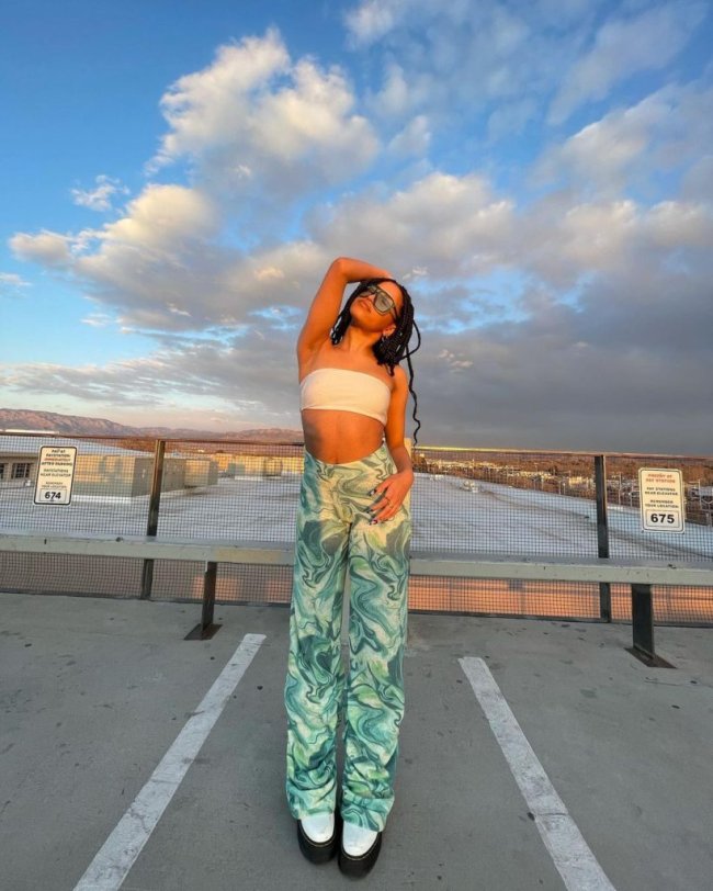 estampas-psicodelicas6-819×1024 Jovem posa em estacionamento com uma mão na cabeça e outra na cintura, ela usa calça verde com estampa psicodélica e top branco.