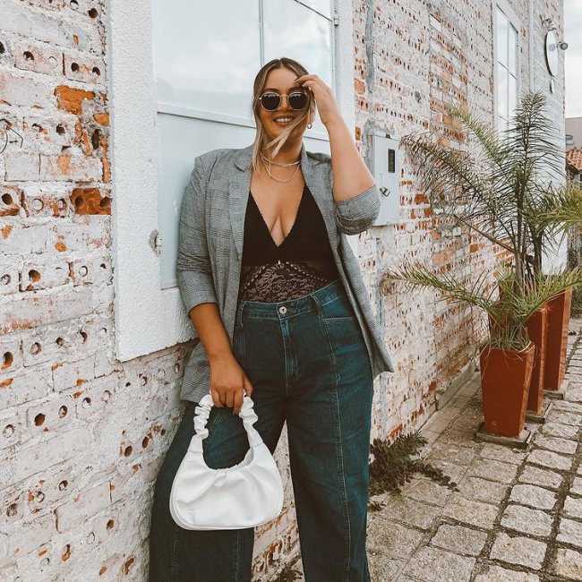 Jovem posando com blazer cinza e body preto, uma de suas mãos está apoiada na cabeça, sua expressão é sorridente e ela está segurando uma bolsa branca.