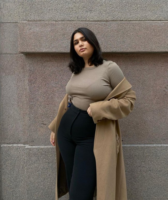 Foto de uma mulher na rua. Ela usa uma camiseta bege, casaco bege e calça skinny preta. Ela olha para a câmera e não sorri.