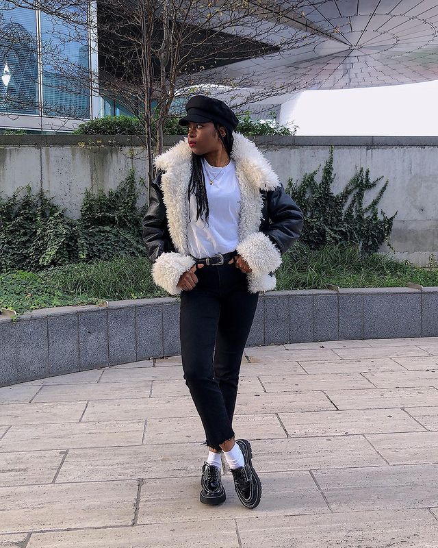 look-calca-skinny-preta Foto de uma mulher em uma calçada. Ela usa uma boina preta, camiseta branca, casaco de pelinho branco e preto, calça skinny preta, cinto preto e mocassim preto. Ela olha para o lado e não sorri.