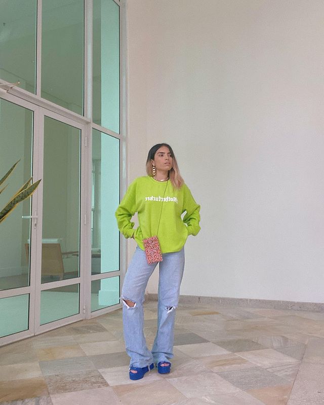 look-verde-com-azul-larissaleall Garota usando moletom verde, calça jeans de lavagem clara rasgada no joelho, sandália de plataforma azul e uma bolsa de miçangas coloridas pendurada no pescoço. Ela está olhando para o lado com as duas mãos na cintura.