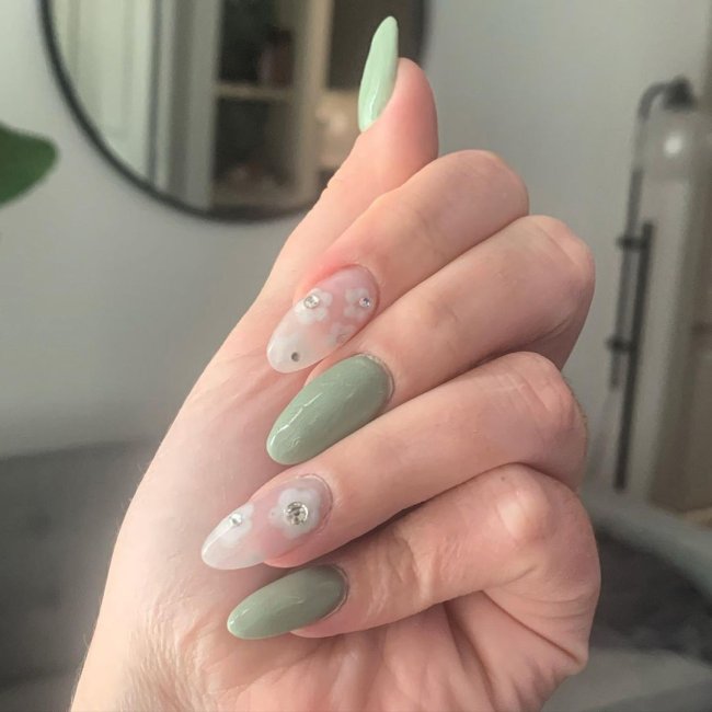 Foto com close nas unhas e tem nail art com desenho de flor branca com bolinha verde.