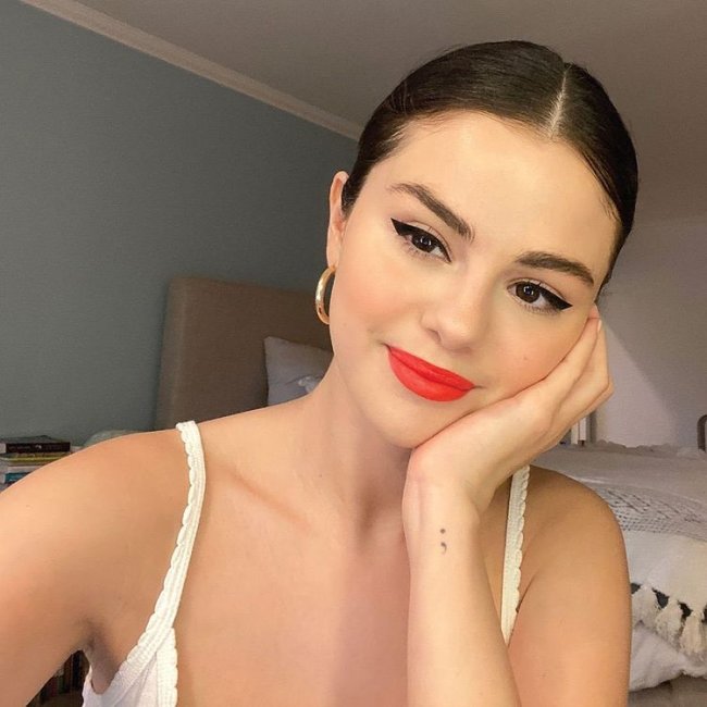 selena-gomez-com-batom-vermelho Selfie da atriz e cantora Selena Gomez. Ela usa uma blusa de alcinha branca, brinco de argola dourado, cabelo preso em um coque e maquiagem com delineado preto e batom vermelho. Ela olha para a câmera, sorri e apoia o rosto na mão esquerda.