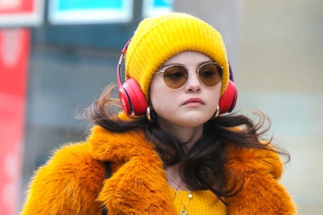 Selena Gomez usando gorro e suéter amarelos, casaco de pelos laranja