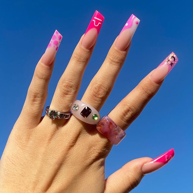 unha-formato-batom Foto de uma mão com o céu como fundo. A mão está com unhas decoradas em formato de batom e três anéis divertidos.