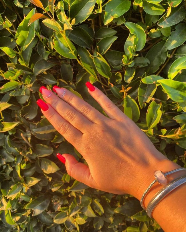 Foto mostrando uma mão apoiada em folhas verdes, nas unhas tem esmalte vermelho com detalhe de strass.